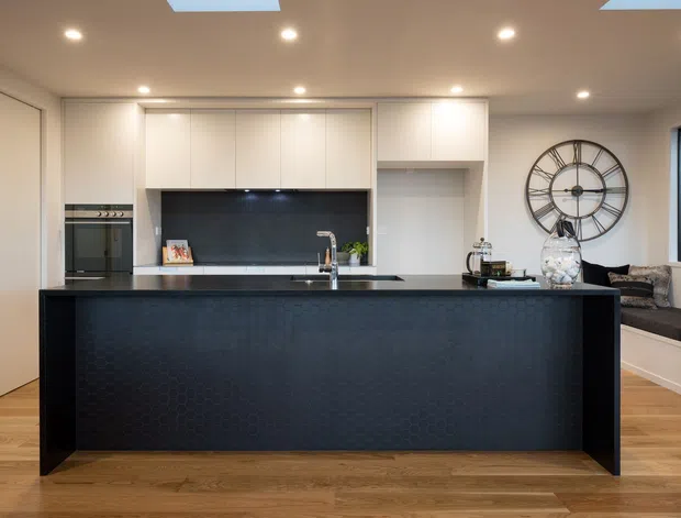 A contemporary open-plan kitchen dominated by a large dark island with an integrated sink and tap. White full-height cabinetry, a built-in oven, dark splashback, recessed ceiling lights, and skylights create a clean, modern fit-out. Timber flooring adds warmth, while a built-in window seat and oversized wall clock are visible in the adjoining living area.