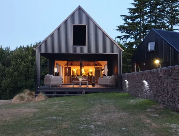Exterior view of a compact gable-roofed dwelling at dusk, featuring vertically ribbed dark cladding, a large open glazed living area, and a raised timber deck. A second matching outbuilding sits behind a gabion-style stone wall, with landscaped lawn and mature trees surrounding the property.