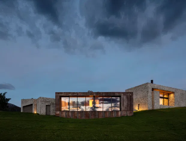 A low, modern residential building set in a broad grassy landscape at dusk. The composition features a long central glazed volume framed in dark weathered metal or timber, flanked by pale textured masonry wings with large warmly lit windows. Dramatic dark clouds dominate the sky, creating a striking contrast with the illuminated interior and rolling lawn.