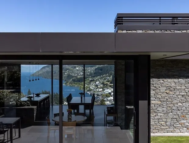Exterior view of a contemporary flat-roofed residence with dark horizontal cladding, extensive floor-to-ceiling glazing, and a stacked stone wall. Through the glazing, a dining and living area is visible, with a dramatic elevated view toward a blue lake and mountainous, wooded terrain. The building has strong horizontal lines, dark framing, and a rooftop or upper-level screened terrace.