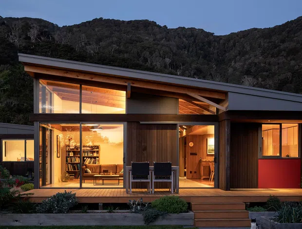 A modern single-storey pavilion-style house at dusk, set against a densely wooded hillside. The dark low-pitched roof and dark cladding frame extensive floor-to-ceiling glazing, revealing a warmly lit timber interior with bookshelves, seating, and timber-lined ceilings. A timber deck, steps, built-in outdoor seating, and landscaped planting occupy the foreground.