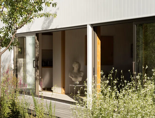 Exterior view of a contemporary single-storey building with a crisp white vertically ribbed facade, broad glazed openings, and a timber deck. The open glass doors reveal a warm, minimalist interior with timber finishes and sculptural decor. Native-style planting and a concrete path frame the building.