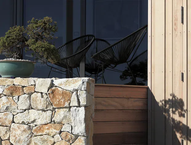 Close-up exterior architectural detail showing a low rustic stone retaining wall, a dark timber-clad gate or panel, and vertical pale timber cladding. Behind the glazing are black sculptural wire lounge chairs, while a large ceramic planter with a bonsai-style tree sits on the stone wall. The composition combines natural stone, timber, dark glazing, and strong sunlight in a refined contemporary facade setting.