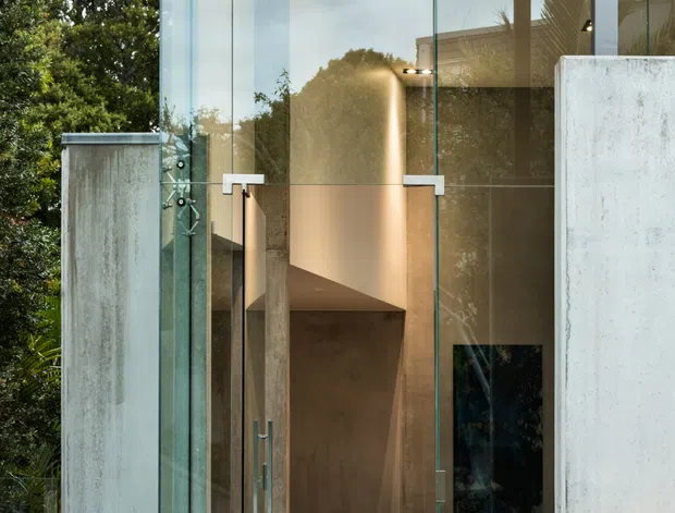 A contemporary concrete-and-glass building entrance viewed from outside. The facade features tall frameless glass doors and glazing, exposed smooth concrete walls and canopy, recessed downlights, and a dark paved entry terrace. Through the transparent entrance, a double-height interior with a dining table and warm lighting is visible, while greenery surrounds the exterior.