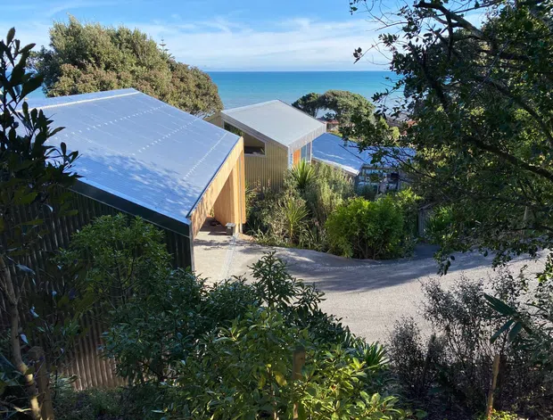 Exterior view of several compact modern buildings stepping down a lush, sloping coastal site, with corrugated metal roofs, timber-clad walls, landscaped planting, and a clear ocean horizon beyond.