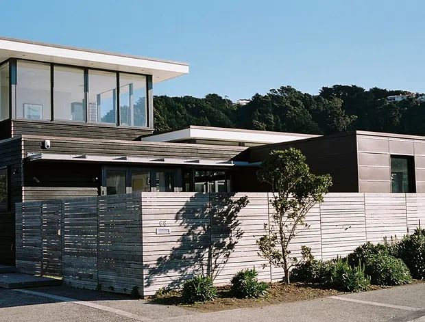 Exterior street view of a contemporary multi-level residential home with dark horizontal cladding, pale timber-toned wall sections, broad floor-to-ceiling upper-level glazing, and several low-profile flat roof planes. A screened front courtyard and landscaped planting sit behind a horizontal slatted fence, with a neighbouring modern dwelling visible at right.