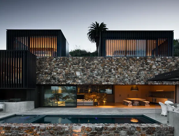 Exterior view of a contemporary multi-level residence at dusk, featuring dark box-like volumes with vertical slatted glazing, a substantial exposed stone retaining wall, expansive glass openings, concrete terraces, and a rectangular swimming pool in the foreground. Warm interior lighting contrasts with the dark facade and surrounding landscape.