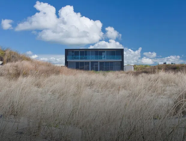 A low, rectilinear modern house is viewed from behind a broad expanse of coastal sand dunes. The building has a dark, box-like facade with extensive floor-to-ceiling glazing and a projecting upper level, set against a vivid blue sky and dune grasses.
