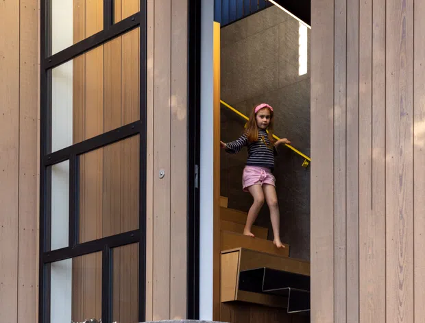 A contemporary residential entry viewed from outside, featuring vertical timber cladding, dark metal-framed glazing, and a partially open doorway revealing a compact timber staircase. The stairwell has dark concrete or textured wall surfaces, a bright yellow handrail, and a blue-toned upper wall. A child is descending the stairs. The foreground includes a cast-concrete boundary wall marked with the number 2015.