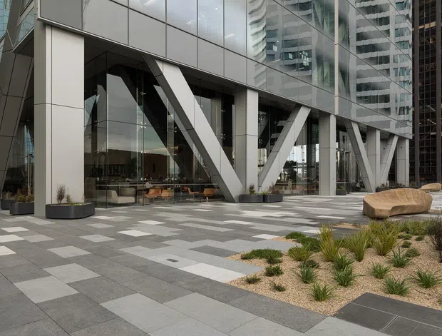 Contemporary commercial building frontage with a glass curtain wall, prominent white structural columns and diagonal bracing, and a broad paved plaza. The forecourt includes geometric grey pavers, low drought-tolerant planting in gravel beds, and sculptural timber seating. High-rise buildings are reflected in and visible through the glazing.