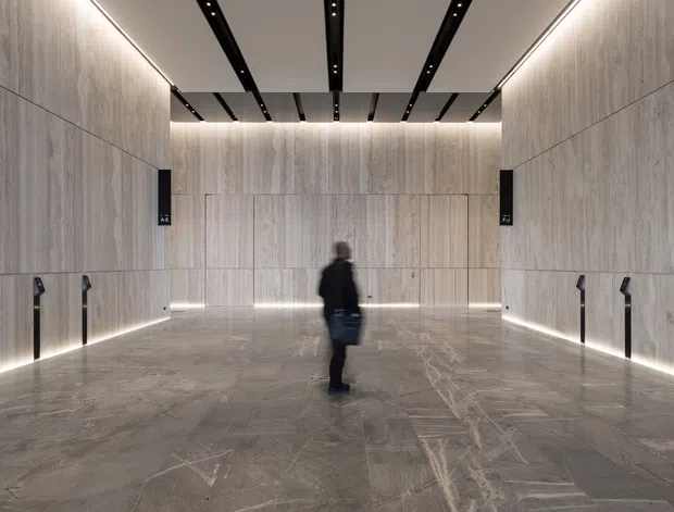 A large, symmetrical contemporary interior corridor or lobby clad in pale stone-look panels, with matching grey stone flooring, linear recessed lighting, dark ceiling tracks, and multiple elevator or door openings along the side walls. A person stands near the centre, emphasizing the generous scale and high ceiling.