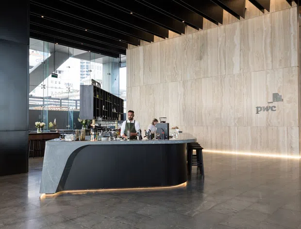 A refined commercial lobby or hospitality reception space featuring a sculptural dark bar or concierge counter, pale stone-clad feature wall with PwC branding, dark exposed ceiling beams, integrated linear lighting, and large glazed windows. The space has a polished, contemporary corporate aesthetic.
