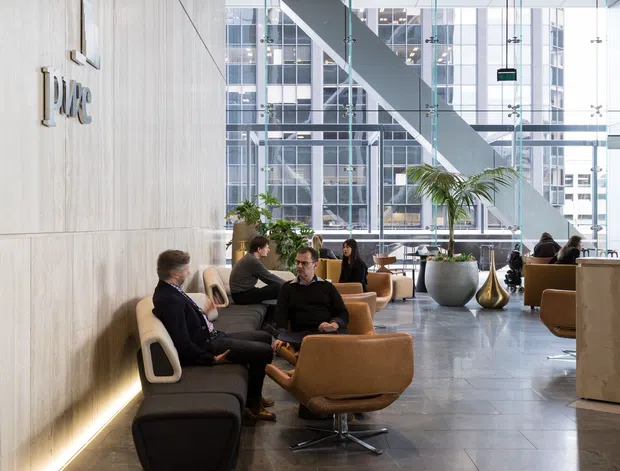 A sophisticated commercial lobby or reception lounge with a large pale timber-panelled feature wall bearing a raised logo, dark exposed linear ceiling battens, extensive floor-to-ceiling glazed curtain walling, and a dramatic diagonal structural element beyond. The space contains clustered tan and dark lounge chairs, small tables, planters, and people using the seating area. Large-format grey stone-look flooring and warm concealed wall lighting create a refined corporate atmosphere.