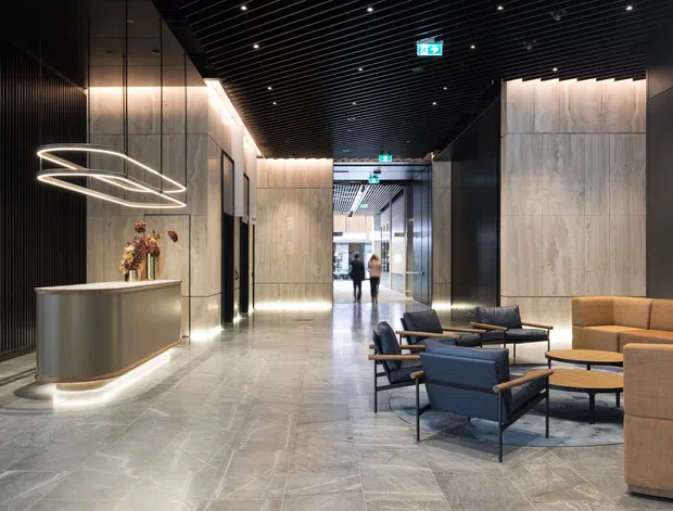 A sophisticated contemporary commercial lobby or reception area with a polished grey stone floor, dark ribbed ceiling and wall detailing, large pale stone wall panels, illuminated linear cove lighting, a sculptural ring pendant, and a curved reception desk. A lounge seating arrangement with upholstered armchairs, sofas, and low coffee tables occupies the foreground.
