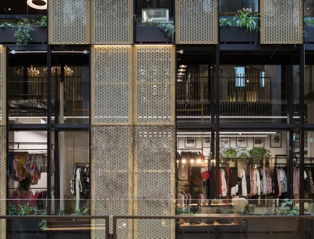 A multi-level contemporary retail interior or shopping arcade viewed across an open atrium. The facade is composed of black-framed glazing, warm brass-toned perforated metal screens, exposed timber slatted beams overhead, and planted balcony edges. Multiple fashion storefronts are visible, including Kate Spade, with pedestrian circulation in the foreground.