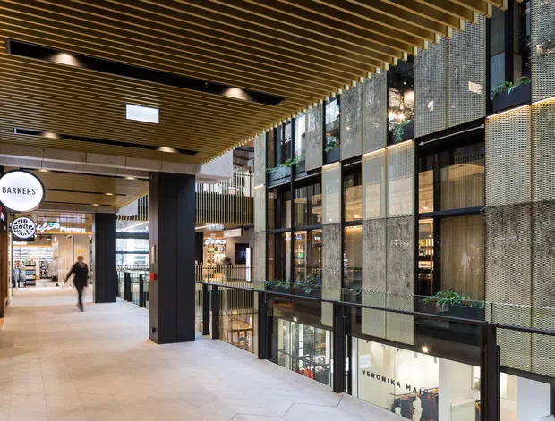 A contemporary multi-level indoor retail and commercial arcade with a wide tiled pedestrian walkway, shopfronts, dark structural columns, planted balcony edges, and a dramatic timber slatted ceiling. The facade on the right combines large glazed panels with perforated metal screens, while branded retail signage and lower-level storefronts are visible.