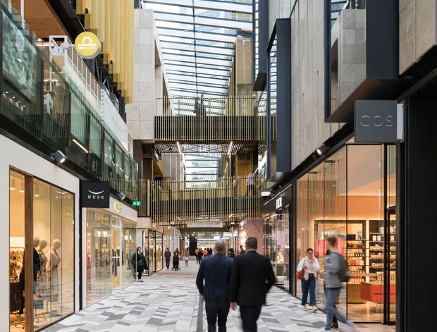 A contemporary multi-level indoor retail arcade or shopping centre with a dramatic glazed skylight roof, exposed structural steel, tall timber-toned vertical screens, concrete and stone walls, and glazed shopfronts lining a wide pedestrian walkway. The floor features a bold geometric chevron pattern in light and dark grey tiles. Shoppers move through the bright, modern commercial space.