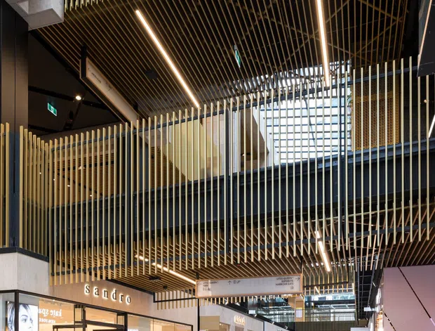 Interior view of a contemporary multi-level shopping centre or commercial retail arcade, featuring layered walkways, dark steel beams, extensive vertical timber-look slat screening, linear integrated lighting, skylights, and polished retail storefronts. Shoppers move through a wide tiled circulation space with visible retail signage and open atrium-style connections between levels.