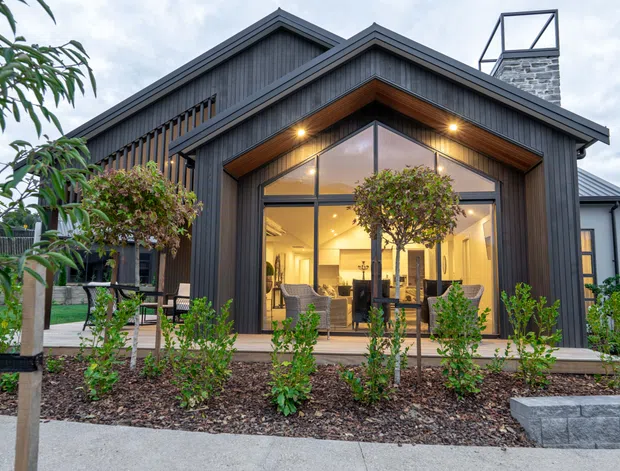 Exterior view of a contemporary gabled house with dark vertical cladding, a steep pitched roof, and a prominent double-height glazed entry or living area beneath a timber-lined gable. Warm interior lighting reveals a furnished room through the glazing. The landscaped foreground includes young trees, shrubs, garden beds, and a low masonry retaining wall.