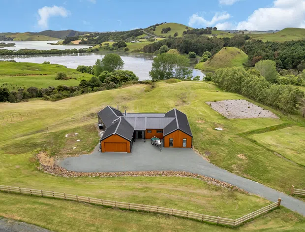 Aerial view of a dark-roofed rural property with orange-brown timber-clad outbuildings, gravel drive, fenced grassy grounds, and a landscaped pond or lake setting amid rolling green hills.
