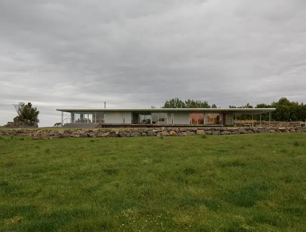 A long, low-slung single-storey modern residence viewed across an open grassy field. The house has extensive glazing, a very shallow flat roof with slender structural supports, and a horizontal composition integrated with a rugged stone retaining edge. The overcast setting and restrained palette create a quiet rural architectural scene.
