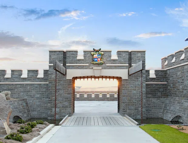 A castle-inspired stone entrance gate viewed from the driveway, featuring crenellated walls, a central arched gateway, raised drawbridge-style timber platform, hanging chains, and a decorative crest beneath the battlement. A flag is visible on the right tower, with landscaped planting and lawn flanking the approach.