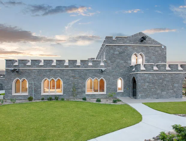 Exterior view of a castle-inspired stone residence with crenellated parapet walls, steep gabled roof sections, pointed-arch windows, and a covered arched entrance. The building is set behind a manicured lawn with curved concrete paths, viewed in warm evening light.