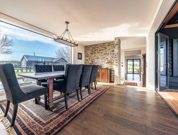 A bright dining area with a long dark wood dining table and upholstered black dining chairs arranged on a patterned rug. The room features wide floor-to-ceiling glazing, warm timber flooring, a rustic exposed-stone feature wall with built-in timber cabinetry, and a large glazed opening to an exterior or covered outdoor space. A linear pendant light hangs above the table, and an adjoining hallway is visible beyond.