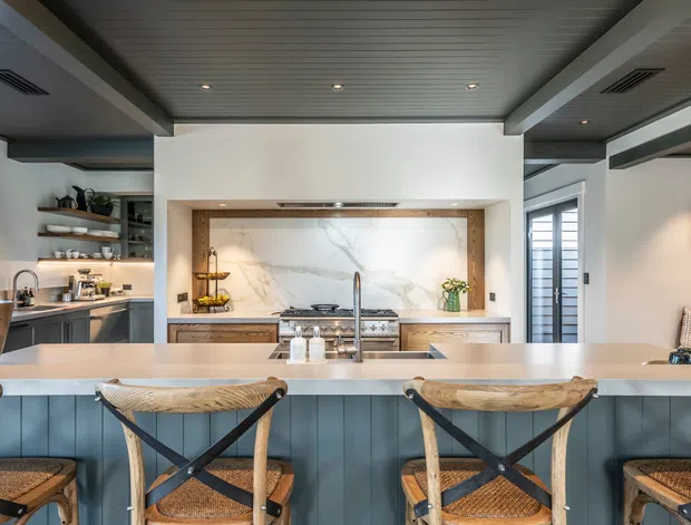Bright, refined kitchen interior viewed from the dining side, featuring a large island with a pale stone benchtop and blue-grey vertical-panel cabinetry, woven timber bar stools, a marble-look splashback, open shelving, integrated appliances, and a dark timber-lined ceiling with recessed lighting. The space has a coastal-contemporary feel with strong contrast between light surfaces and dark blue cabinetry.