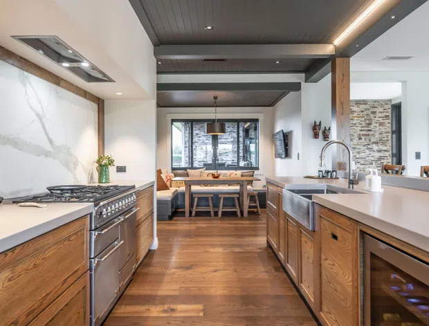 A spacious, open-plan kitchen with a central island, timber cabinetry, stainless-steel appliances, and light stone benchtops. The kitchen opens to a dining and living area, with exposed timber beams and dark timber ceiling panels. A large marble-look splashback, pendant lighting, and a textured stone feature wall add visual interest.