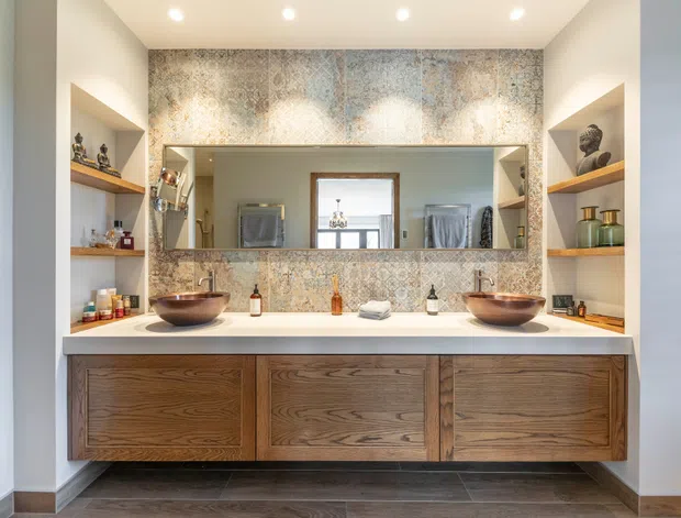 A spacious contemporary double-vanity bathroom with a floating dark timber vanity, two vessel basins, a pale stone benchtop, and a wide wall-to-wall mirror. Patterned decorative tile forms the backsplash and extends to the ceiling, while open timber display shelves flank the vanity. Recessed ceiling lights provide bright, even illumination.