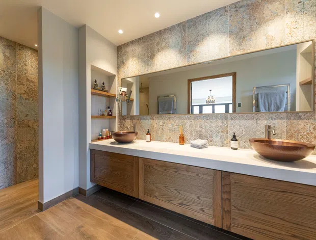 A spacious contemporary bathroom featuring a long double vanity with warm wood-grain cabinetry, a white solid-surface benchtop, and two vessel basins. The walls are clad in richly patterned blue-grey and beige stone or stone-look tile, complemented by timber-look flooring. A large horizontal mirror reflects a window and additional bathroom details, while recessed ceiling lights provide even illumination.