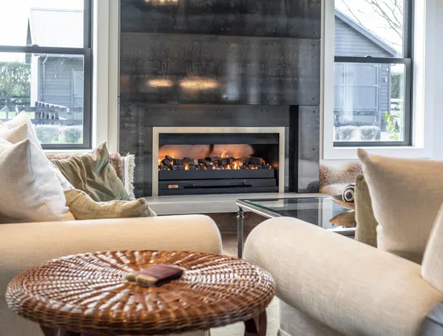 Bright contemporary living room centered on a modern built-in fireplace with a dark textured feature wall. Large windows flank the fireplace, while neutral upholstered sofas and armchairs surround a glass coffee table and woven round ottoman.