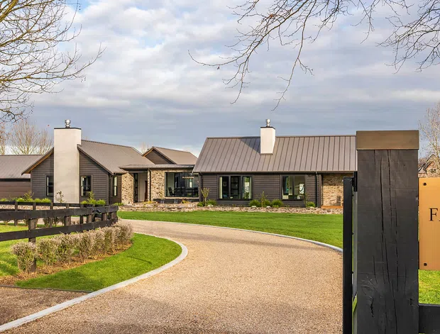 A wide exterior view of a low-slung residential property approached by a curved gravel driveway. The home has dark horizontal cladding, stone feature walls, multiple pitched metal roof sections, and prominent chimneys. A dark timber entrance gate in the foreground carries a brass-toned sign reading “FAIRWOOD 119,” with manicured lawns and mature bare trees surrounding the property.