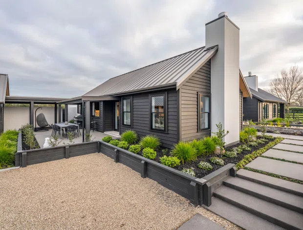 Exterior view of a contemporary single-storey home with dark horizontal cladding, a pitched metal roof, prominent white chimney volumes, and extensive landscaped garden beds. The property includes a gravel forecourt, stepped paved walkway, raised dark-edged planters, outdoor dining area, and covered outdoor connection between building volumes.