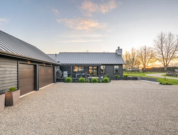 Exterior view of a single-storey dark-clad residential property with a large gravel driveway, attached or adjacent double garage, metal pitched roofs, and landscaped garden areas. The low horizontal building forms, dark weatherboard-style cladding, and rural open setting are visible in warm late-day light.