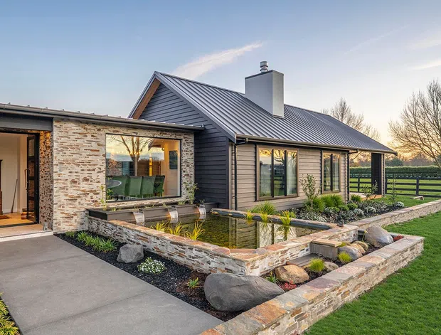 A single-storey contemporary rural home with dark horizontal cladding, a pitched standing-seam metal roof, large windows, and a pale stone-clad extension. The front entry and living area overlook a landscaped rectangular pond with stone edging, boulders, planted garden beds, and a lawn beyond.