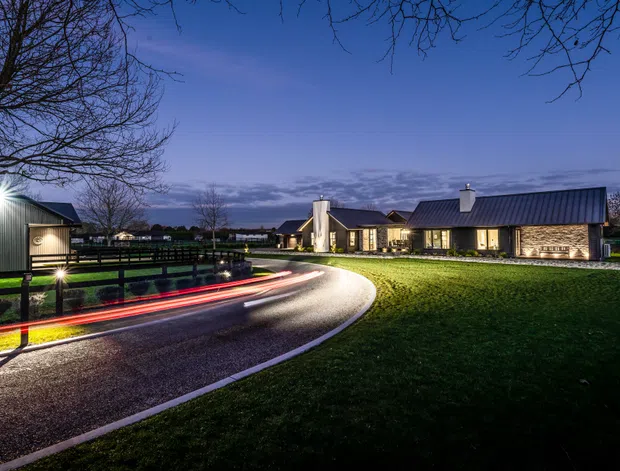 Twilight exterior view of a low, contemporary rural property arranged around a curved driveway. The buildings feature dark standing-seam pitched roofs, light rendered walls, large illuminated windows, and a connected garage or outbuilding. A timber fence, manicured lawn, and long-exposure vehicle light trails emphasize the approach to the residence.
