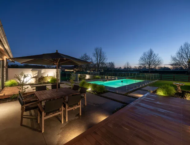 Dusk view of a landscaped outdoor entertaining area with a timber-clad house, covered dining patio, large umbrella, timber decking, illuminated swimming pool, lawn and garden planting. The scene has open rural or suburban views beyond the property and a warm, inviting atmosphere.