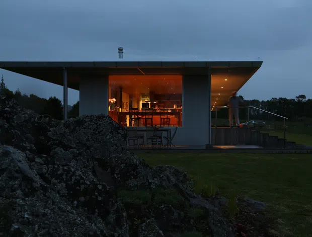 A compact modern pavilion set among rocky natural landscape, photographed at dusk. The building has a dramatic low-pitched flat roof with broad overhangs, dark vertical cladding or structural panels, extensive floor-to-ceiling glazing, and a warmly illuminated open-plan interior. A covered terrace with outdoor furniture extends along the right side, while exposed rock formations and lawn surround the structure.