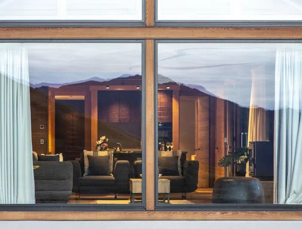 Exterior-facing view through a large timber-framed glazed facade, showing a warmly lit contemporary living room with dark upholstered seating, timber wall finishes, curtains, a fireplace-like illuminated feature, and a plant. Mountainous terrain is visible beyond and reflected in the glazing.
