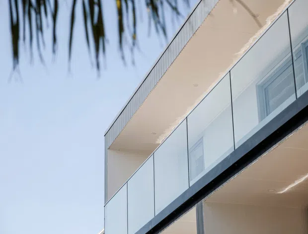 Close-up exterior view of a contemporary multi-level building, focused on projecting balconies with clear glass balustrades, light-coloured soffits, large glazed openings, and clean rectilinear lines. Palm fronds and blue sky appear in the background, suggesting a warm coastal or subtropical setting, though the architecture is the main subject.