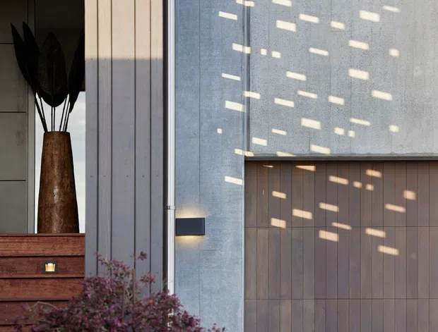 Close-up of a contemporary building facade featuring a tall grey concrete or rendered wall, vertical timber-clad entry or garage door, slim dark window and door framing, and a minimalist wall light. A timber console and tall plant are visible through adjacent glazing, while patterned sunlight creates rectangular highlights across the facade.