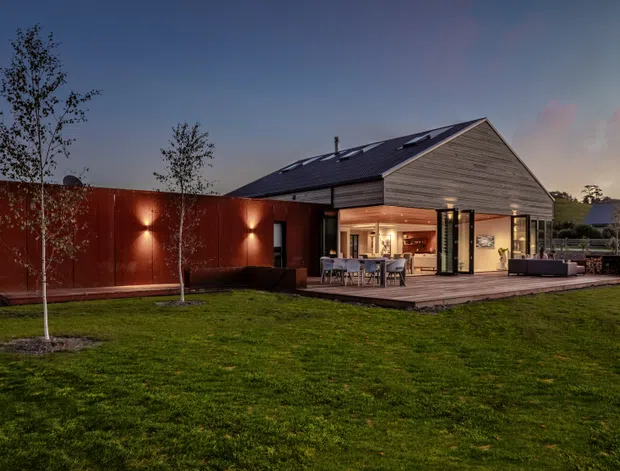 Twilight exterior view of a contemporary single-storey home set in a landscaped lawn. The house combines a steep gabled roof with dark roofing, horizontal timber-look cladding on the gable, dark glazed openings, and a contrasting rust-red metal-clad wing. A timber deck extends from the open-plan living and dining area, which is visible through the large glazed doors.