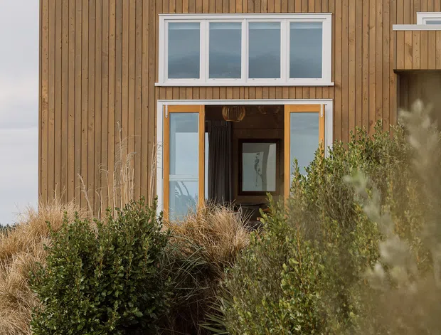 Exterior view of a modern two-storey building with vertical timber cladding, white-framed windows and glazed sliding doors. The entrance is partly concealed by native-style landscaping and ornamental grasses, with a timber fence and pathway in the foreground. A solar panel and chimney are visible above the roofline.