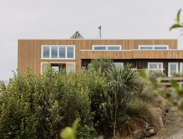 Exterior view of a contemporary, box-like multi-level home clad in warm vertical timber boards. The facade features large horizontal windows, a projecting upper-level volume, a covered balcony or terrace with timber balustrade, and a simple low-pitched or flat roofline. Dense coastal-style planting partially screens the lower facade.