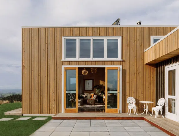 Exterior view of a modern two-storey timber-clad home opening onto a paved terrace. The facade features vertical warm wood boards, large white-framed windows, glazed timber doors, and a projecting upper-level form. Outdoor white metal seating is arranged beside the entrance, with landscaped lawn and a broad rural landscape visible beyond.