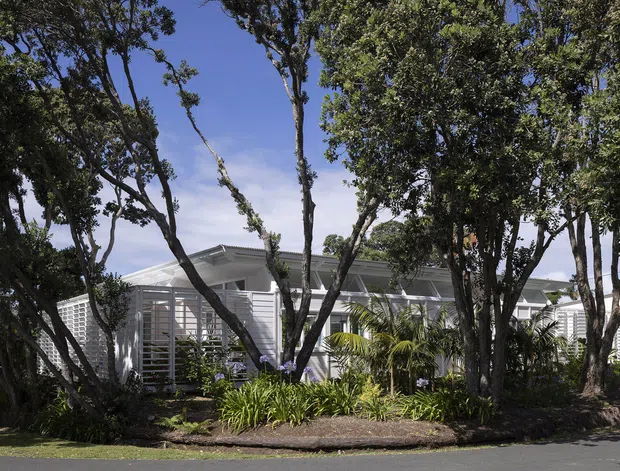 Exterior view of a low, light-coloured contemporary residence partly screened by mature trees and tropical landscaping. The building features broad horizontal forms, white louvred or slatted privacy screens, extensive glazing, and a low-pitched roofline.
