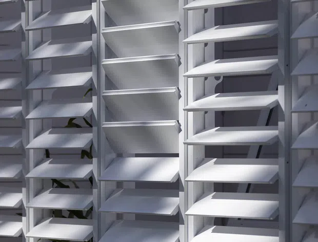 Close-up view of a large modular louvred screen or shading facade composed of repeated white or light-grey horizontal angled slats set within vertical frames. Strong sunlight creates alternating highlights and shadows, with a few small green plants visible behind the screen. The image focuses on the architectural facade system rather than an interior room.