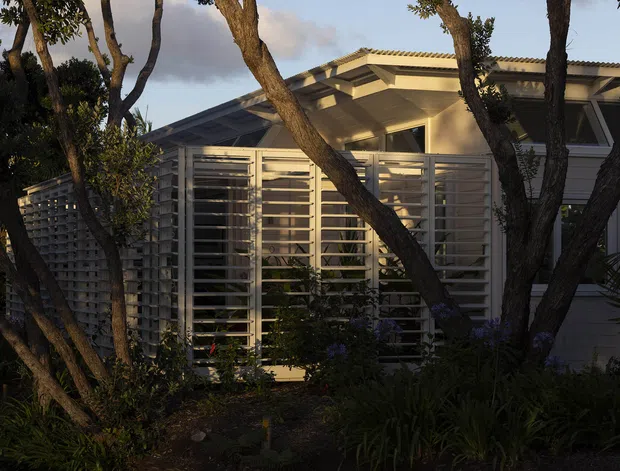 Exterior view of a low-rise residential building partially screened by mature trees and dense planting. The light-coloured facade features extensive horizontal louvres and a projecting covered entry or verandah, with a shallow pitched corrugated roof and shaded glazing behind the screens.